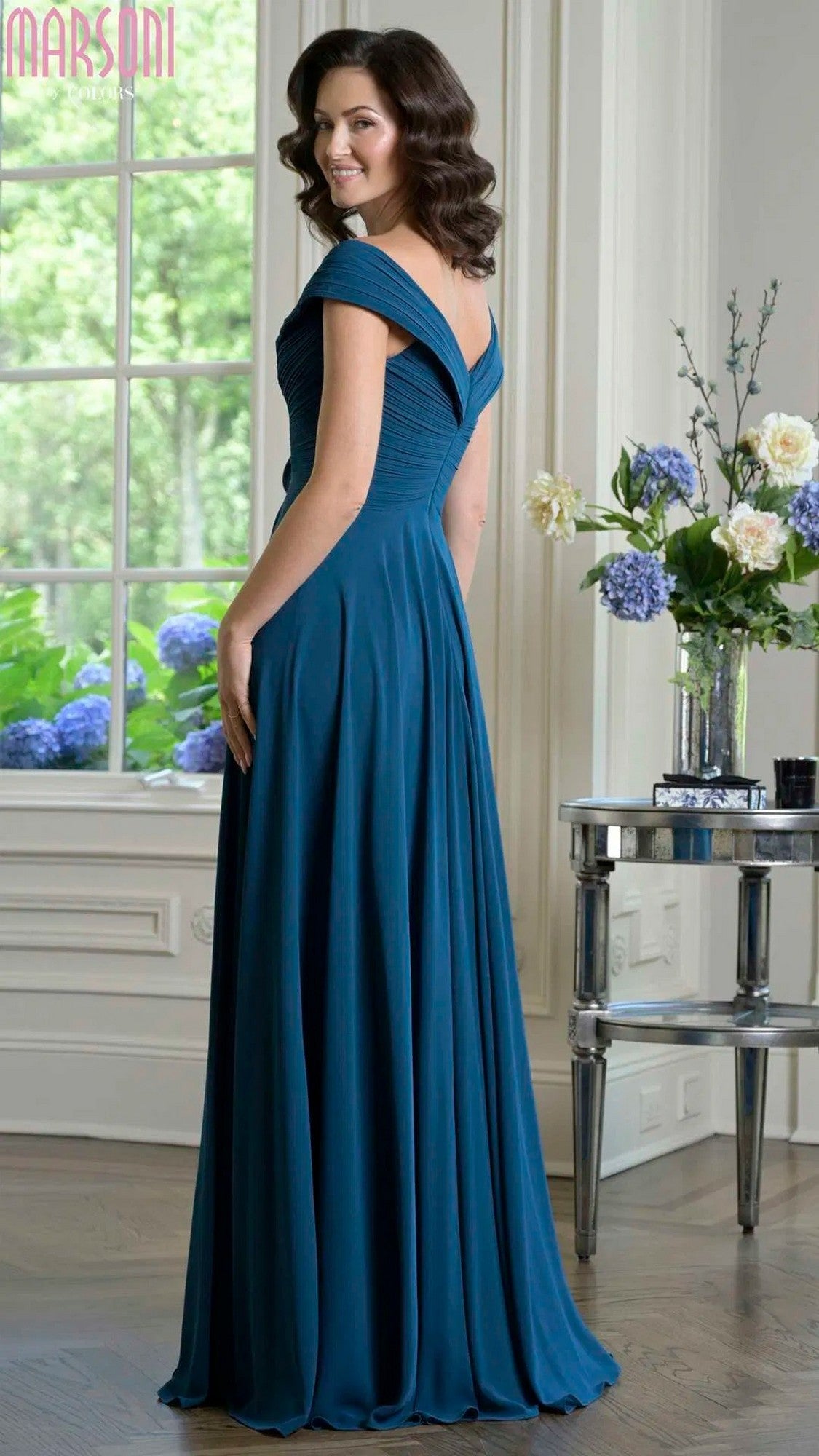 Woman in a Marsoni M333 Long Formal Dress in teal stands indoors by a window and table with flowers, looking over her shoulder and smiling.